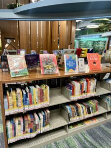 A library book display featuring a sign for the Word Collectors Club, surrounded by colorful children's books. The sign stands out among titles like Actual Size and Perfectly Norman, inviting young readers to explore and collect words.
